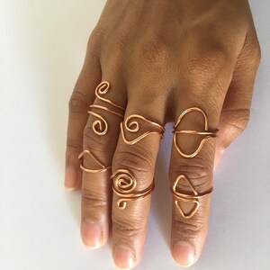 May include: A set of eight copper wire rings in various shapes and sizes. The rings are worn on the fingers of a hand.