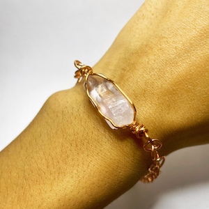 Könnte beinhalten: Ein goldfarbenes, drahtgewickeltes Armband mit einem Anhänger aus klarem Quarzkristall.