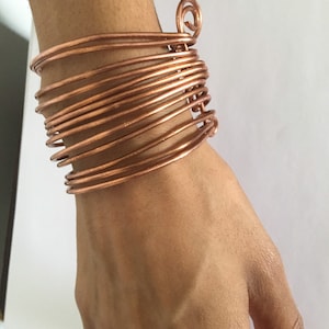 Könnte beinhalten: Ein Armband aus Kupferdraht mit einem Spiraldesign. Das Armband besteht aus mehreren Drahtlagen, wodurch es dick und robust wirkt.