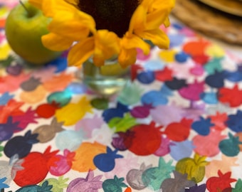May include: A white table runner with a colorful pattern of painted fruit. The fruit is in various shades of red, orange, yellow, green, blue, and purple. A sunflower is in the foreground.