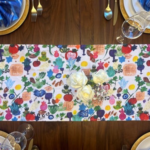 May include: A white table runner with a colorful, hand-painted pattern featuring images of pomegranates, apples, grapes, and other symbols of the Jewish holiday of Rosh Hashanah. The runner is decorated with the Hebrew word "shalom" in a repeating pattern.