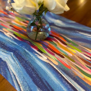 May include: A white floral arrangement in a clear glass vase sits on a blue and white table runner with colorful abstract brushstrokes.
