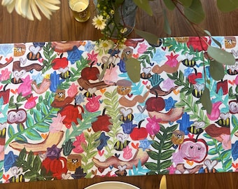 Könnte beinhalten: Ein weißer Tischläufer mit einem farbenfrohen, fantasievollen Muster aus Früchten, Blumen und Bienen. Der Läufer liegt auf einem Holztisch mit einem Gedeck, das einen weißen Teller, goldenes Besteck und eine zu einer Schleife gefaltete Serviette umfasst.