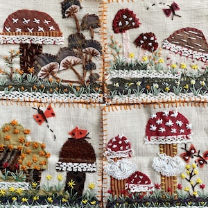May include: Four handmade fabric squares with embroidered mushroom designs. Each square features different mushroom shapes and colors, including red, white, brown, and green. The squares are stitched together with a white lace trim and orange stitching.