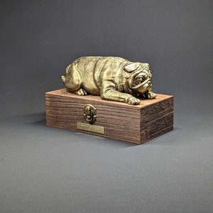 May include: A gold-colored pug figurine resting on a wooden box with a brass latch and a nameplate that reads "Dioby".