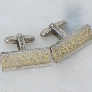 May include: A pair of rectangular silver cufflinks with a textured, light-colored center. The cufflinks have a hinged bar for securing to a shirt cuff. The design is simple and elegant, suitable for formal wear.