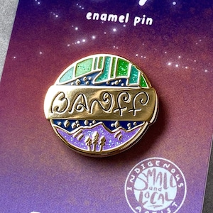 May include: Gold enamel pin with a purple, green, and blue mountain scene and the word "Banff" in gold lettering. The pin is on a purple and pink background with the text "Art by Jimmy" and "enamel pin".