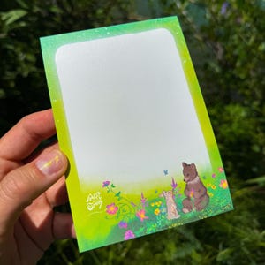 May include: A notepad with a green and white gradient border. The bottom of the notepad features a whimsical illustration of a brown bear, a rabbit, and colorful flowers. The text "Art Ginny" is in the lower left corner.