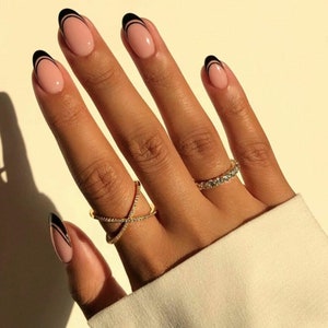 May include: Close-up of a hand with nude-colored nails featuring a black double-line design. The hand is wearing two gold rings with small clear stones.