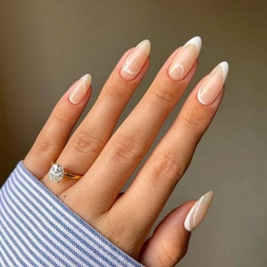 May include: Close-up of a hand with almond-shaped nails painted with a nude polish and white abstract designs. The nails are long and have a slight curve. The hand is wearing a gold ring with a large diamond.