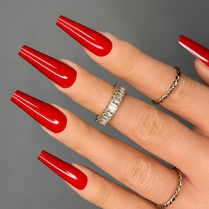 May include: Close-up of a hand with long, red, coffin-shaped nails. The hand is wearing a thin gold band with clear, rectangular stones.
