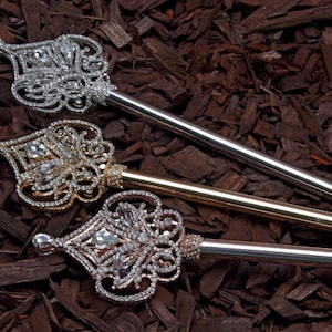May include: Three ornate jeweled scepters with gold, silver, and rose gold finishes. The scepters are lying on a bed of brown wood chips.