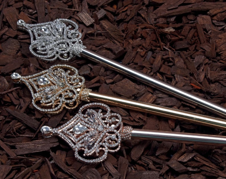 May include: Three ornate jeweled scepters with gold, silver, and rose gold finishes. The scepters are lying on a bed of brown wood chips.