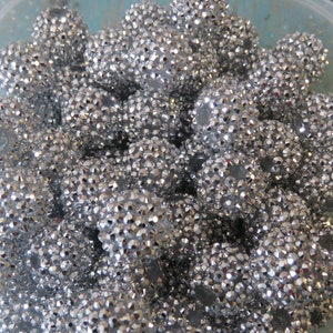May include: A close-up of a container filled with silver rhinestone balls. The balls are all the same size and are arranged in a random pattern. The container is made of clear plastic and has a white background.