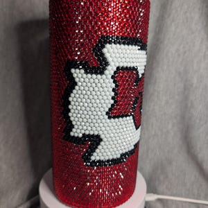 K&ouml;nnte beinhalten: Ein roter Becher, bedeckt mit funkelnden roten Strasssteinen. Der Becher hat ein wei&szlig;es und schwarzes Design. Das Design ist ein stilisiertes "K" mit wei&szlig;em Hintergrund und schwarzer Umrandung. Der Becher ist zylindrisch und steht auf einer wei&szlig;en Basis.