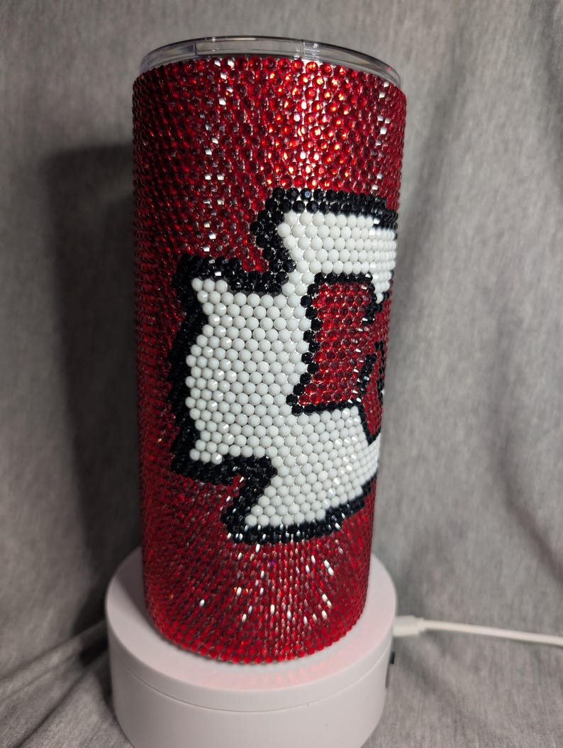 K&ouml;nnte beinhalten: Ein roter Becher, bedeckt mit funkelnden roten Strasssteinen. Der Becher hat ein wei&szlig;es und schwarzes Design. Das Design ist ein stilisiertes "K" mit wei&szlig;em Hintergrund und schwarzer Umrandung. Der Becher ist zylindrisch und steht auf einer wei&szlig;en Basis.