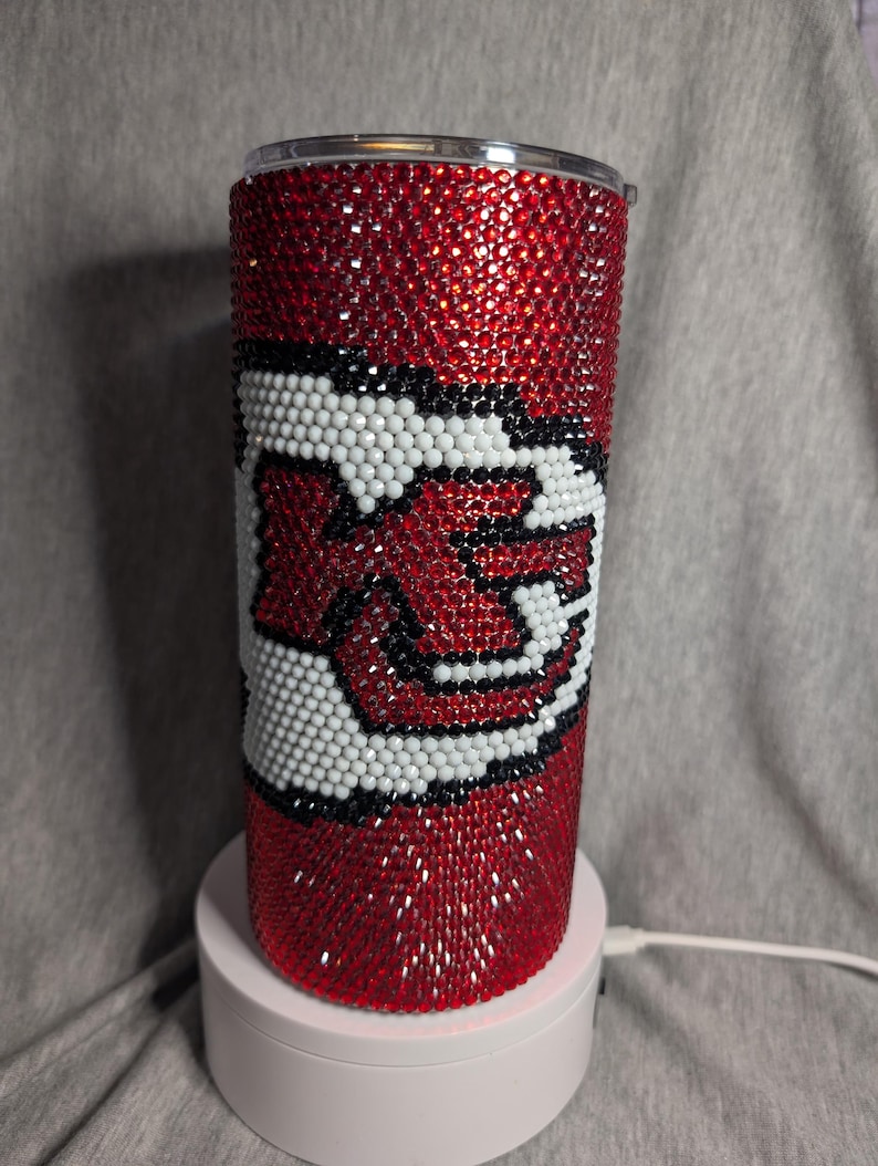 K&ouml;nnte beinhalten: Ein roter, mit Strasssteinen besetzter Becher mit einem Teamlogo in Wei&szlig;, Schwarz und Rot. Der Becher ist mit kleinen, funkelnden Strasssteinen bedeckt. Das Teamlogo ist prominent auf der Vorderseite des Bechers zu sehen.