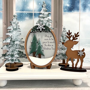 May include: A white wooden base with three wooden Christmas decorations. The decorations include a reindeer, a snow-covered tree, and a round ornament with two trees inside and the text "May you never be too grown to search the skies on Christmas Eve".