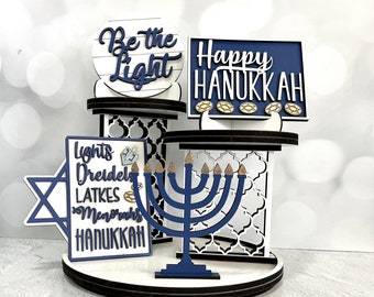 May include: A white tiered stand with blue and gold accents features a menorah, a star of David, and three signs that read "Be the Light", "Happy Hanukkah", and "Lights, Dreidels, Latkes, Menorahs, Hanukkah".