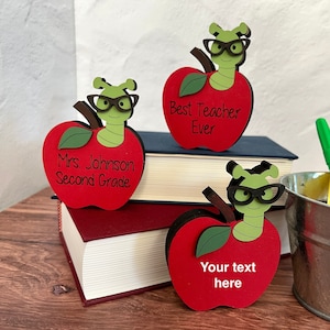 May include: Three wooden apple ornaments with a worm wearing glasses on top. The apples are red with green leaves. The text on the apples reads "Best Teacher Ever", "Mrs. Johnson Second Grade", and "Your text here".