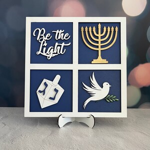 May include: A white wooden frame with four squares. Each square has a different image: a menorah, a dreidel, a dove with an olive branch, and the words "Be the Light".