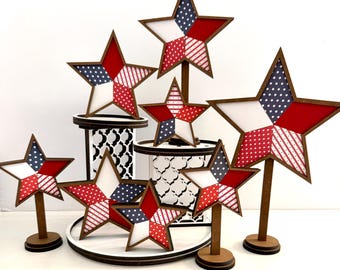 May include: A collection of patriotic star decorations in red, white, and blue. The stars have a wooden frame and are displayed on stands and tiered platforms. The stars feature various patterns, including polka dots and stripes, and are ideal for Independence Day.