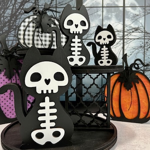 May include: Black and white skeleton cat figurines with a pumpkin and a black and white checkered pumpkin on a tiered stand. The stand is on a black round tray.