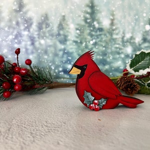 Pode incluir: Uma figura de p&aacute;ssaro cardeal vermelho com um ramo de azevinho e bagas vermelhas. A figura &eacute; feita de madeira e tem um acabamento pintado. O fundo &eacute; uma imagem desfocada de uma floresta nevada.