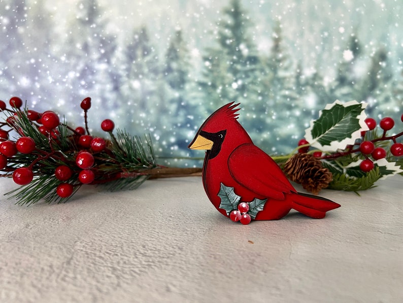 Pode incluir: Uma figura de p&aacute;ssaro cardeal vermelho com um ramo de azevinho e bagas vermelhas. A figura &eacute; feita de madeira e tem um acabamento pintado. O fundo &eacute; uma imagem desfocada de uma floresta nevada.