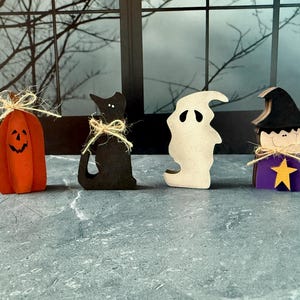 May include: A collection of Halloween-themed wooden decorations. Includes an orange pumpkin with a carved face, a black cat, a white ghost, and a witch figure with a purple base and a yellow star. Each figure is tied with twine.