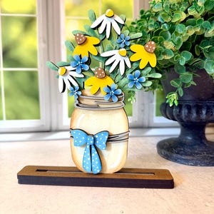 May include: A wooden mason jar decoration with a bouquet of daisies. The flowers are white, yellow, and blue, with green leaves. The jar is cream-colored with a blue polka-dot bow. It sits on a brown wooden base.