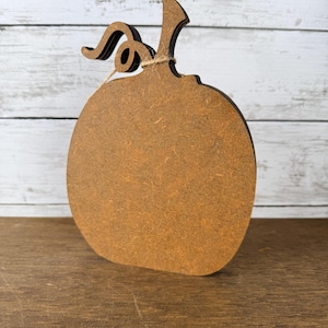 May include: A flat, pumpkin-shaped cutout made of brown composite wood material. The object features a curved stem and a small, decorative piece of twine tied around the base of the stem. The edges of the pumpkin are dark brown in colour.