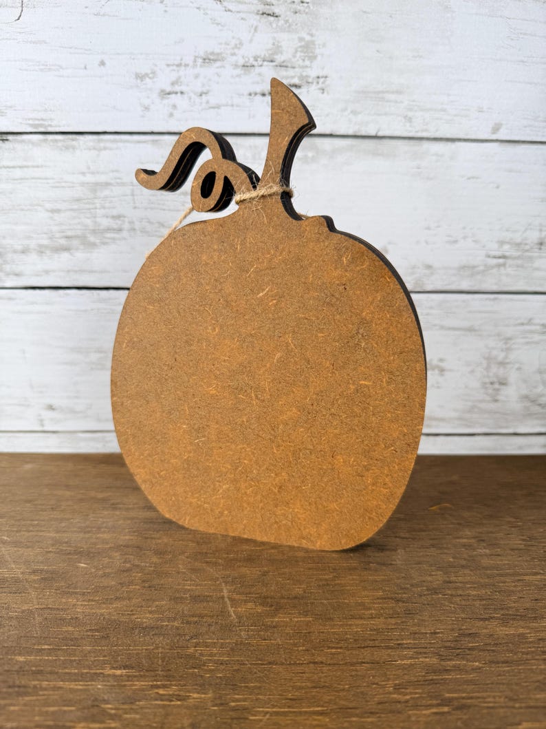 May include: A flat, pumpkin-shaped cutout made of brown composite wood material. The object features a curved stem and a small, decorative piece of twine tied around the base of the stem. The edges of the pumpkin are dark brown in colour.