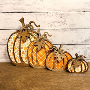 May include: A set of four graduated wooden pumpkin decorations featuring laser-cut outlines and stems with attached twine bows. Each pumpkin has a unique patterned insert, including autumn leaves, orange plaid, white polka dots, and floral motifs.