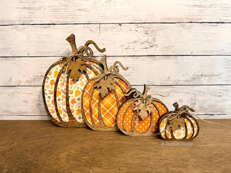 May include: A set of four graduated wooden pumpkin decorations featuring laser-cut outlines and stems with attached twine bows. Each pumpkin has a unique patterned insert, including autumn leaves, orange plaid, white polka dots, and floral motifs.