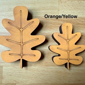 May include: Two orange-yellow wooden oak leaf cutouts of different sizes. Each leaf features a central vein and branching side veins etched into the surface. The leaves have a smooth, flat finish and are shaped with rounded lobes.