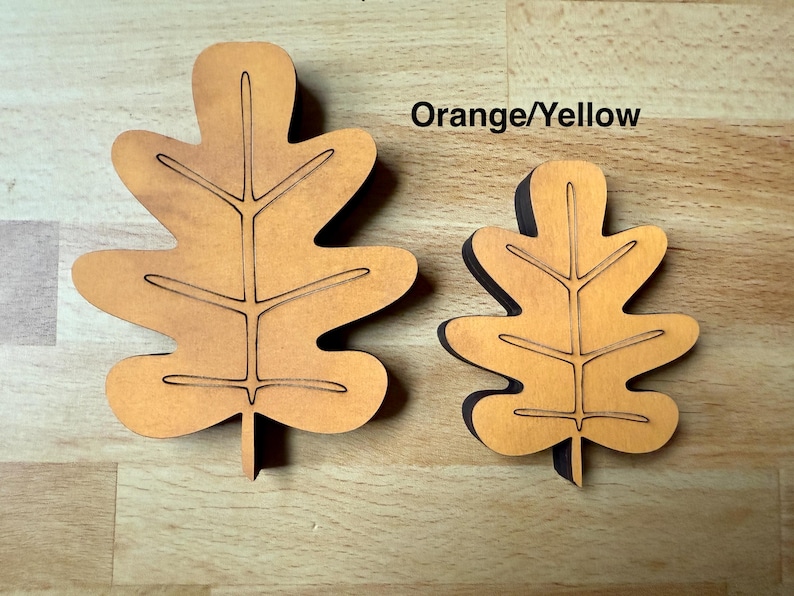 May include: Two orange-yellow wooden oak leaf cutouts of different sizes. Each leaf features a central vein and branching side veins etched into the surface. The leaves have a smooth, flat finish and are shaped with rounded lobes.