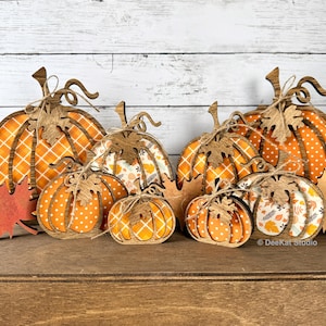 May include: A collection of wooden pumpkin-shaped decorations in various sizes, featuring orange and white patterns including plaid, polka dots, and autumn leaves. Each pumpkin is accented with a small wooden leaf and twine bow.