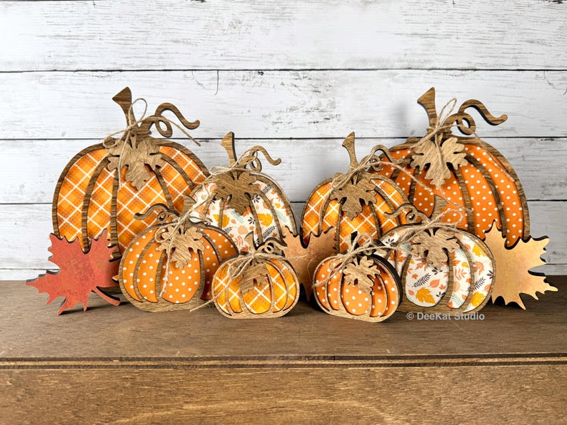 May include: A collection of wooden pumpkin-shaped decorations in various sizes, featuring orange and white patterns including plaid, polka dots, and autumn leaves. Each pumpkin is accented with a small wooden leaf and twine bow.