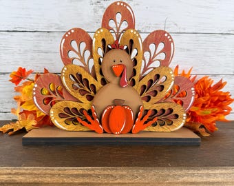May include: A layered wooden turkey decoration featuring a tan body, orange feet, and a red wattle. The tail feathers are cut in intricate patterns with shades of brown, orange, and yellow. A small orange pumpkin sits in front of the turkey.