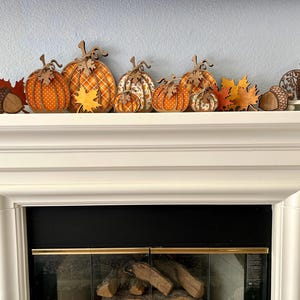May include: A collection of wooden autumn-themed decorations featuring orange and yellow pumpkins with polka dot and plaid patterns, brown acorns, maple leaves, and a small bird silhouette, all arranged in a row on a white fireplace mantel.
