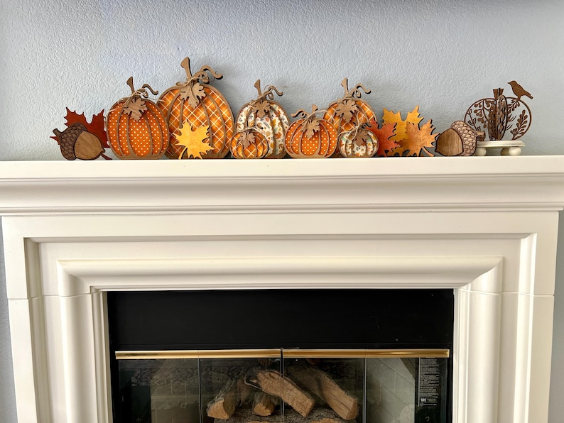 May include: A collection of wooden autumn-themed decorations featuring orange and yellow pumpkins with polka dot and plaid patterns, brown acorns, maple leaves, and a small bird silhouette, all arranged in a row on a white fireplace mantel.