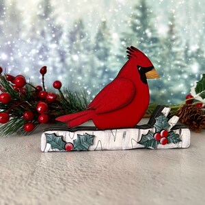 Pode incluir: Um p&aacute;ssaro cardeal vermelho empoleirado em um tronco de b&eacute;tula branco com folhas de azevinho verdes e bagas vermelhas. O fundo &eacute; uma cena de inverno com neve.
