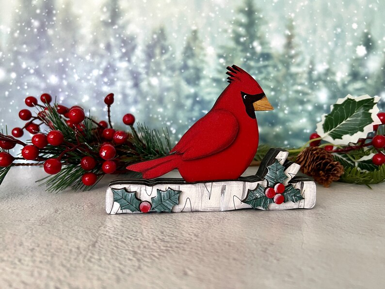 Pode incluir: Um p&aacute;ssaro cardeal vermelho empoleirado em um tronco de b&eacute;tula branco com folhas de azevinho verdes e bagas vermelhas. O fundo &eacute; uma cena de inverno com neve.
