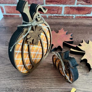 May include: Two layered wooden pumpkin decorations featuring an orange and white plaid pattern, black edges, and a jute twine bow with a small wooden leaf accent. Two separate wooden maple leaf cutouts in orange and brown tones are also included.