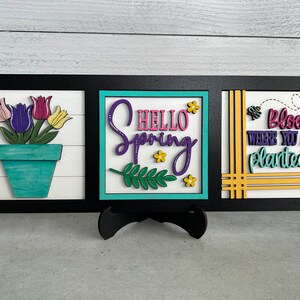 K&ouml;nnte beinhalten: Drei Holzschilder mit Fr&uuml;hlingsmotiv. Das linke Schild zeigt eine Topfpflanze mit lila, rosa und gelben Bl&uuml;ten. Das mittlere Schild sagt "Hello Spring" in lila und rosa Schrift. Das rechte Schild sagt "Bloom Where You Are Planted" in lila und gr&uuml;ner Schrift.