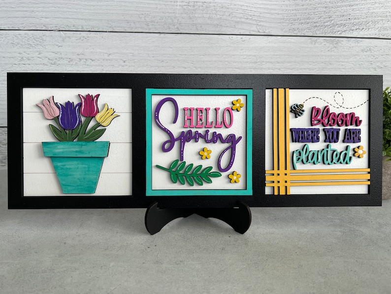 K&ouml;nnte beinhalten: Drei Holzschilder mit Fr&uuml;hlingsmotiv. Das linke Schild zeigt eine Topfpflanze mit lila, rosa und gelben Bl&uuml;ten. Das mittlere Schild sagt "Hello Spring" in lila und rosa Schrift. Das rechte Schild sagt "Bloom Where You Are Planted" in lila und gr&uuml;ner Schrift.