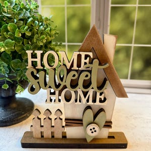 May include: A wooden house-shaped tabletop sign featuring the text HOME SWEET HOME in layered lettering. The design includes a small picket fence, a sage green heart accent with a white button, and a brown roof, all mounted on a flat base.
