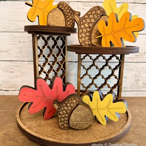May include: A tiered wooden display stand with two levels featuring intricate lattice cutouts. The stand is decorated with several wooden cutouts of autumn leaves in shades of orange, red, and yellow, along with detailed wooden acorn shapes.