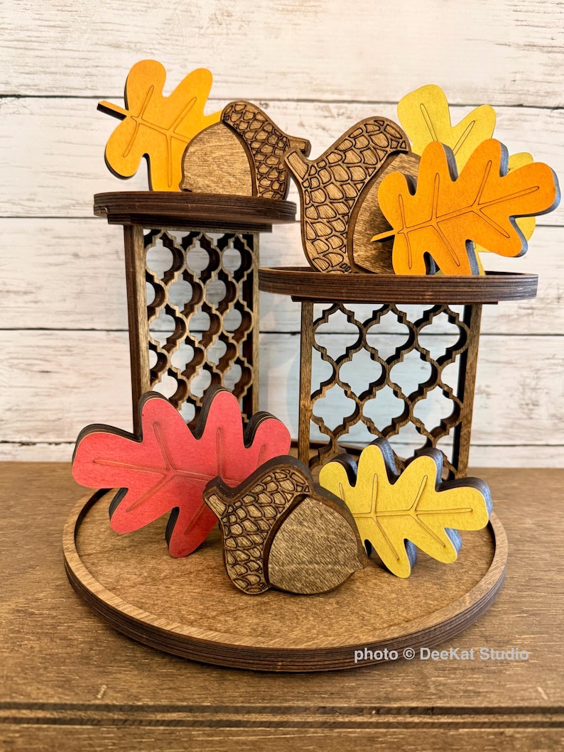May include: A tiered wooden display stand with two levels featuring intricate lattice cutouts. The stand is decorated with several wooden cutouts of autumn leaves in shades of orange, red, and yellow, along with detailed wooden acorn shapes.
